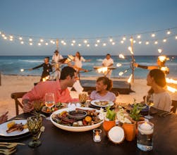Beachfront dinner at sunset with a table full of delicious food and entertainment near the shoreline.