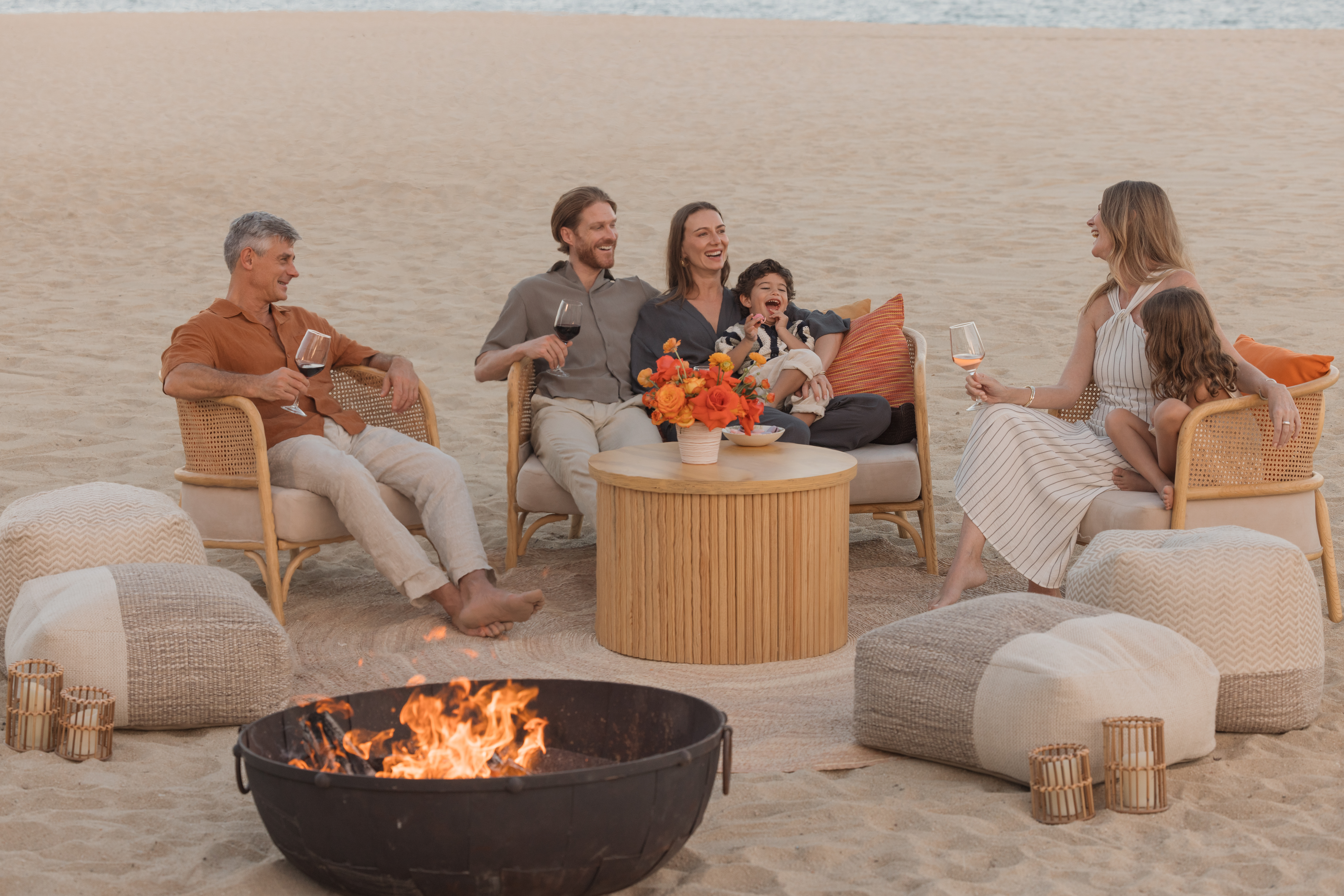 Family sitting around a beachside fire pit, enjoying drinks in a relaxed lounge setup.